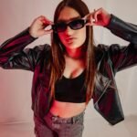 Trendy young woman with sunglasses and leather jacket posing confidently in a studio setting.